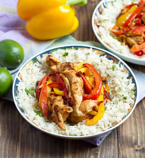 strips of seasoned chicken, red, and yellow bell peppers in bowl on top white rice with cilantro and black pepper; another bowl with same ingredient in background; 2 limes to left of bowl and yellow bell pepper behind bowl; light lavender cloth under bowl and produce