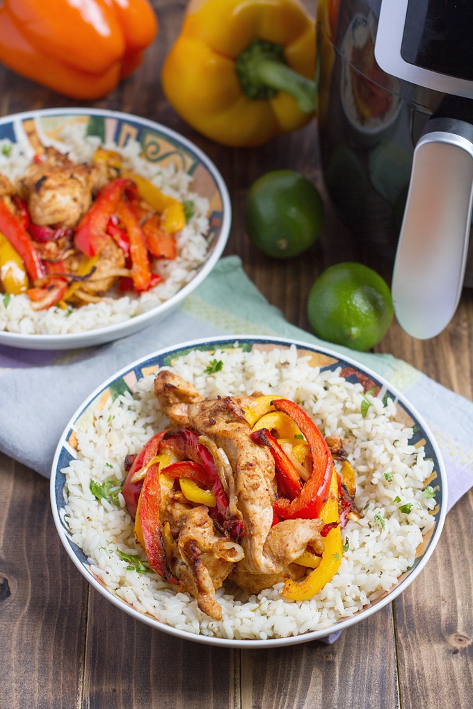 strips of seasoned chicken, red, and yellow bell peppers in bowl on top white rice with cilantro and black pepper; another bowl with same ingredient in background; 2 limes to left of bowl and orange and yellow bell peppers behind bowl; light lavender cloth under bowl; air fryer in top right corner