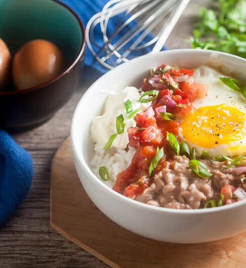 bowl with rice, dollop of sour cream, salsa, sunnyside up egg, refried beans, and chopped green onion garnish on light brown platter; cilantro and whick behind bowl; dark brown bowl with robin egg blue interior with 2 brown eggs in it and a bright blue cloth behind the bowl
