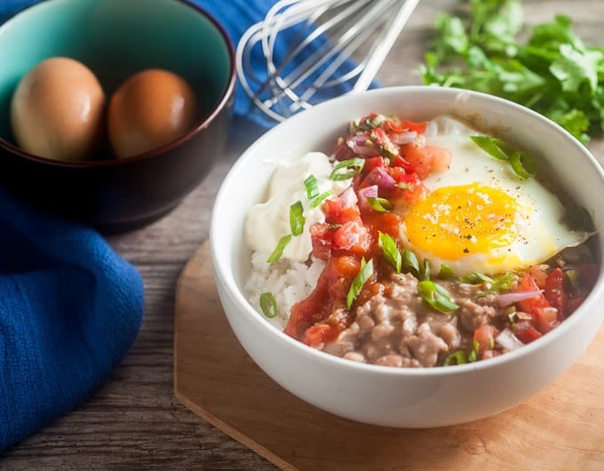 bowl with rice, dollop of sour cream, salsa, sunnyside up egg, refried beans, and chopped green onion garnish on light brown platter; cilantro and whick behind bowl; dark brown bowl with robin egg blue interior with 2 brown eggs in it and a bright blue cloth behind the bowl
