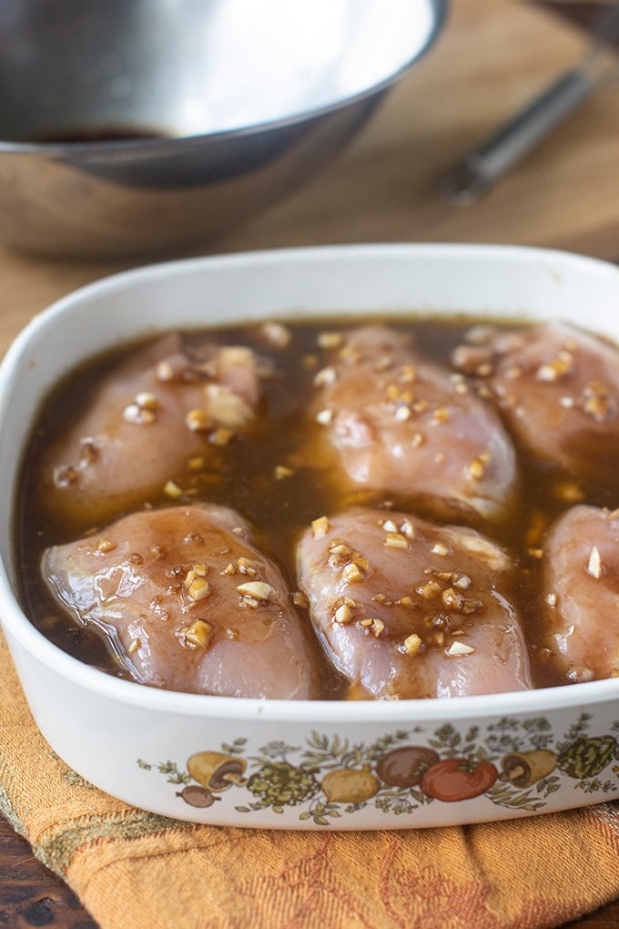chicken thighs in small white casserole dish with handles; sitting in brown marinade liquid with minced garlic; metal bowl and knife in background