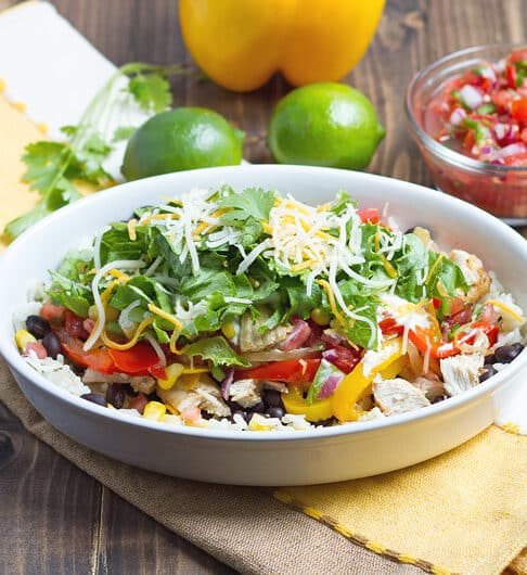oblong white bowl with rice with cubes of chicken, slices of red and yellow bell pepper, lettuce, and white and orange shredded cheese; white, yellow, and beige striped cloth under bowl; cilantro, lime, yellow bell pepper, and fresh salsa in condiment bowl
