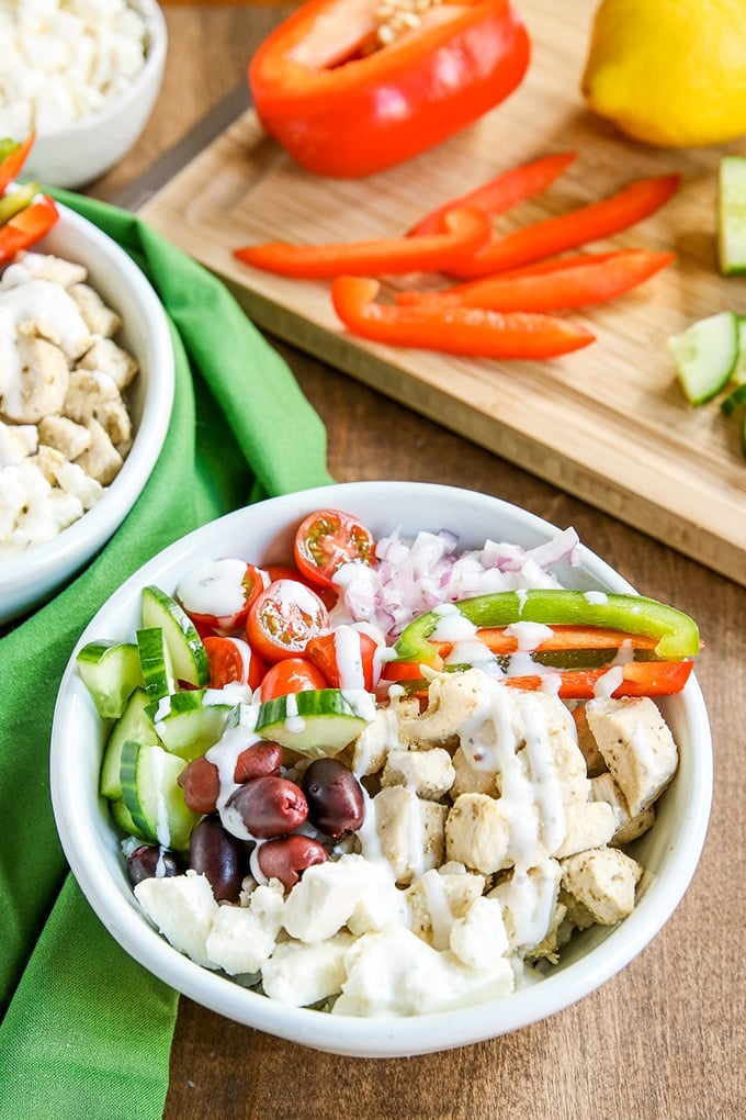 Greek Rice Bowls white bowl with rice, chicken, kalamata olives, cucumber, green and red bell peppers, halved cherry tomatoes with white sauce drizzled over; cutting board in background with orange bell pepper, lemon, and cucumber; medium colored green cloth behind bowl