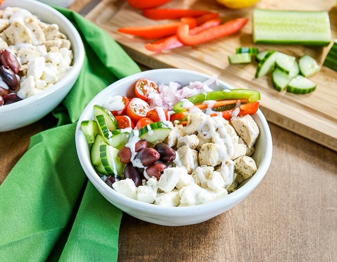 white bowl with rice, chicken, kalamata olives, cucumber, green and red bell peppers, halved cherry tomatoes with white sauce drizzled over; cutting board in background with orange bell pepper, lemon, and cucumber; medium colored green cloth behind bowl