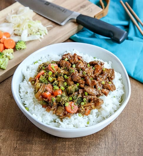white bowl with rice topped with beef, carrots, broccoli, and onion mixture; light blue cloth in background on right with chopsticks on it; back left has a small cutting board with chef's knife, chopped broccoli, carrots, and onion