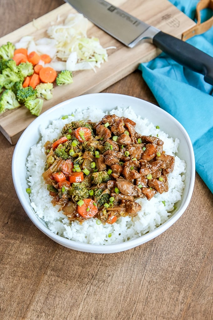 white bowl with rice topped with beef, carrots, broccoli, and onion mixture; light blue cloth in background on right; back left has a small cutting board with chef's knife, chopped broccoli, carrots, and onion