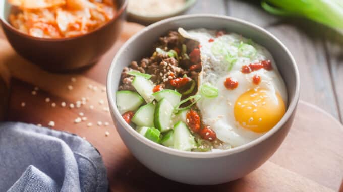 white bowl with rice topped with cucumber, beef, green onion garnish, sesame seeds, sunnyside up egg, and drizzle of sriracha; in background, blurred out condiment bowl, green onion,and bowl with something orange in it; light blue cloth in front left corner; sesame seeds on table