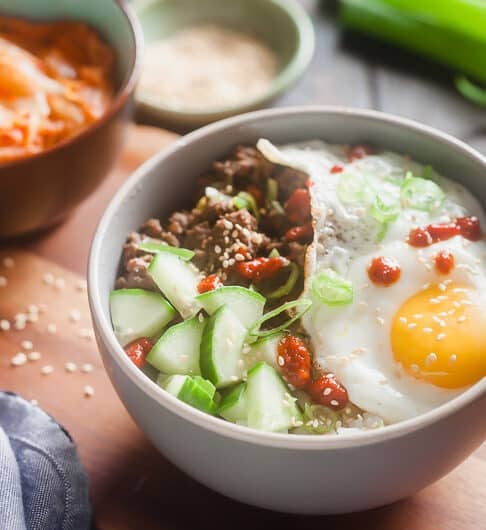 white bowl with rice topped with cucumber, beef, green onion garnish, sesame seeds, sunnyside up egg, and drizzle of sriracha; in background, blurred out condiment bowl, green onion,and bowl with something orange in it; light blue cloth in front left corner; sesame seeds on table