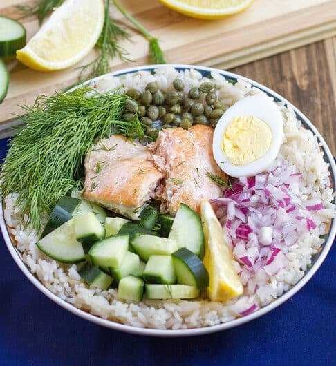 rice in bowl topped with cucumber, salmon, fresh dill, capers, half hard boiled egg, chopped red onion, and lemon wedge; dark blue cloth under bowl; cutting board in background with cucumber rounds, lemon, wedges, and dill