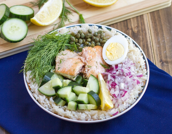 rice in bowl topped with cucumber, salmon, fresh dill, capers, half hard boiled egg, chopped red onion, and lemon wedge; dark blue cloth under bowl; cutting board in background with cucumber rounds, lemon, wedges, and dill