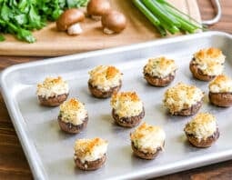 silver baking tray with stuffed mushrooms on it; cutting board in background with parsley, crimini mushrooms, and green onion top