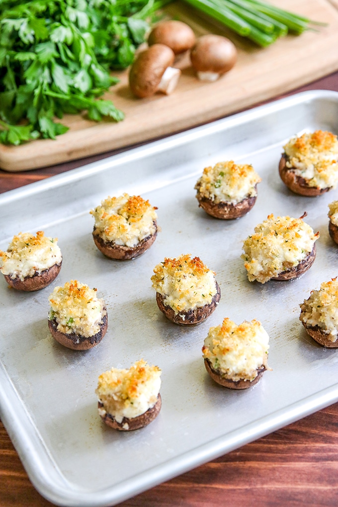 silver baking tray with stuffed mushrooms on it; cutting board in background with parsley, crimini mushrooms, and green onion top