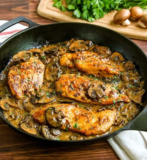 cast iron pan with chicken breast in mushroom sauce with green garnish; white cloth with red stripe underneath; cutting board in background with crimini mushrooms and parsley garnish