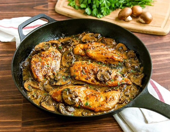 cast iron pan with chicken breast in mushroom sauce with green garnish; white cloth with red stripe underneath; cutting board in background with crimini mushrooms and parsley garnish