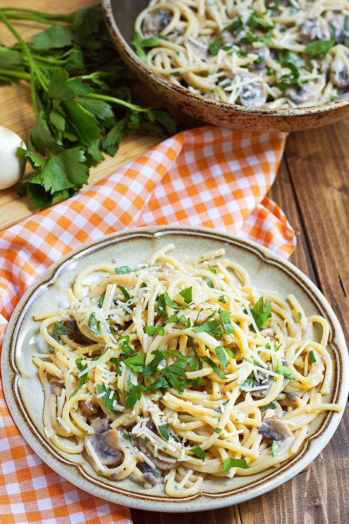 spaghetti with mushrooms, parsley, and shredded cheese; orange and white gingham cloth to left of plate; fresh parsley bunch in back right corner of picture