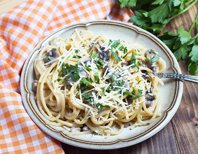 spaghetti with mushrooms, parsley, and shredded cheese; orange and white gingham cloth to left of plate; fresh parsley bunch in back right corner of picture