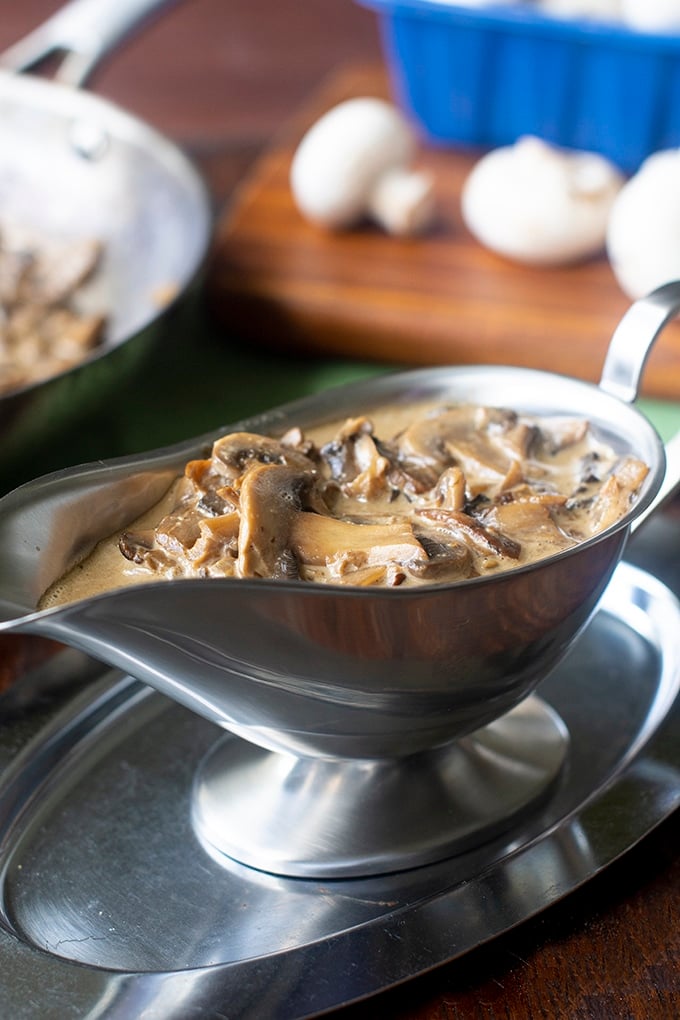 stainless steel gravy boat with mushroom gravy in it; brown cutting board with white mushrooms and blue mushroom container on in background and edge of pan with gravy in it in back left