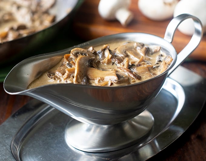 stainless steel gravy boat with mushroom gravy in it; brown cutting board with white mushrooms on in background and edge of pan with gravy in it in back left