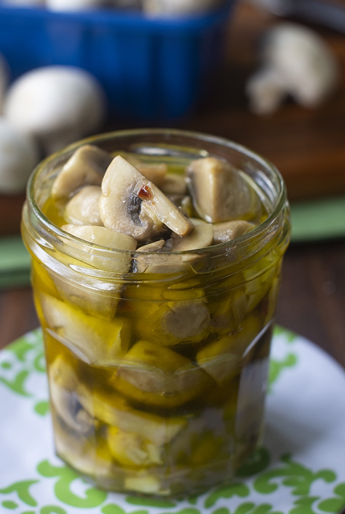 jelly jar with mushroom pieces marinating in oil; white and green plate underneath with blurred out white mushrooms and blue mushroom container in background