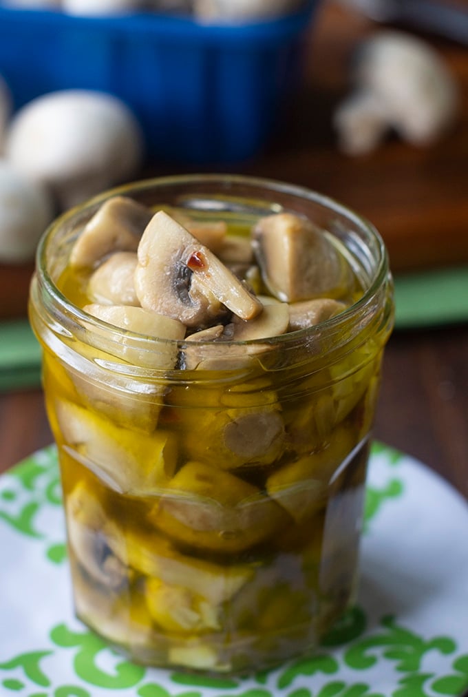 glass jar with mushrooms in oil sitting on small white plate with green design; cutting board in background with white mushrooms on it and blue plastic mushroom container in background