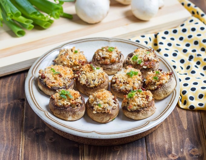 white plate with brown strip around edge with stuffed mushrooms topped with breadcrumbs and green onion garnish; ellow cloth to right with black polka dots; background has a light brown striped cutting board with a bit of green onion tops showing