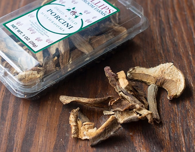 Package of dried mushrooms dried mushrooms on dark wood table with clear plastic container in background that has white label with green edge that says porcini