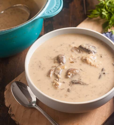 creamy mushroom soup in white bowl with spoon to left and natural wood board under bowl; medium blue cloth to right; parsley garnish in background; back left has partial of a le creuset teal pot with mushroom soup and ladle in it