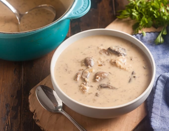 creamy mushroom soup in white bowl with spoon to left and natural wood board under bowl; medium blue cloth to right; parsley garnish in background; back left has partial of a le creuset teal pot with mushroom soup and ladle in it