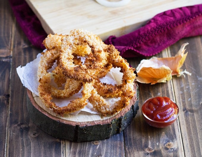 onion rings piled on a round wooden board with paper towel on top; little condiment bowl with ketchup in it to right; onion to left; burgundy cloth in the background with cutting board on it