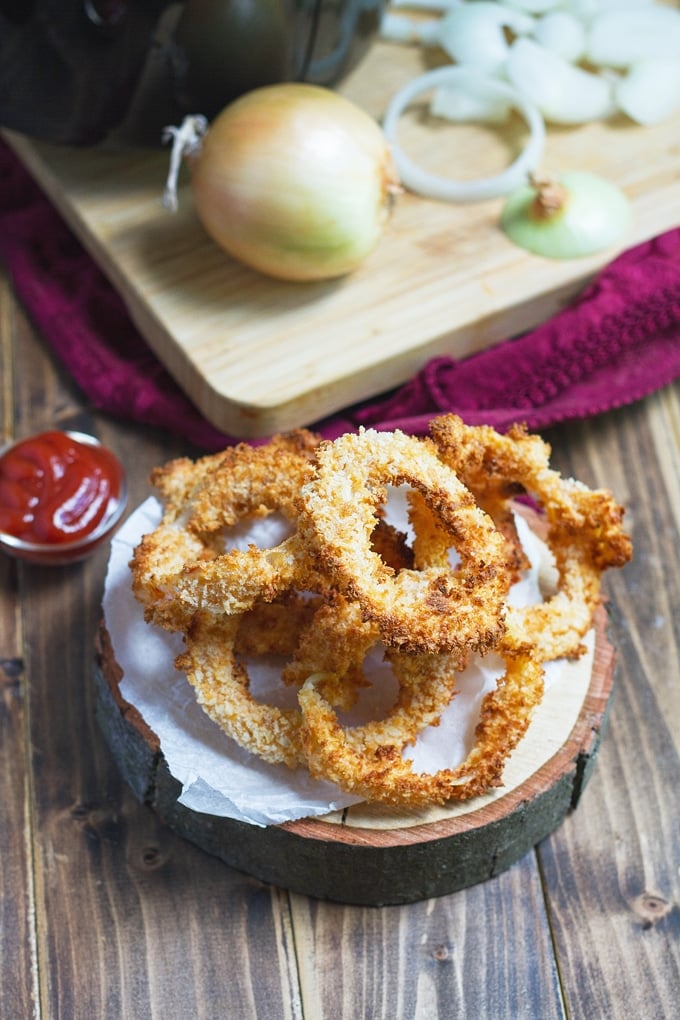 Air Fryer Onion Rings onion rings piled on a round wooden board with paper towel on top; little condiment bowl with ketchup in it to right; onion to left; burgundy cloth in the background with cutting board on it with an onion and sliced onion rings