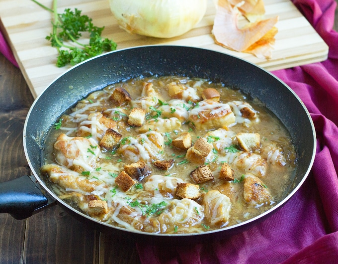 frying pan with pieces of chicken and croutons in french onion soup topped with melted cheese and fresh parsley; plum colored clothto right of pan and cutting board in background with fresh parsley and onion
