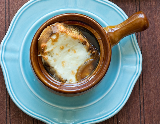 light blue plate with a brown long handled crock of french onion soup with a piece of toasted baguette and melted cheese