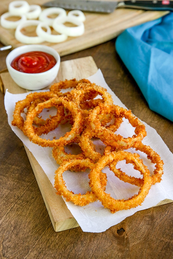 fried onion rings on a wooden board with a paper towel under it; white condiment bowl on background with ketchup; light blue cloth in back right corner; cutting board in background with uncooked onion rings and knife