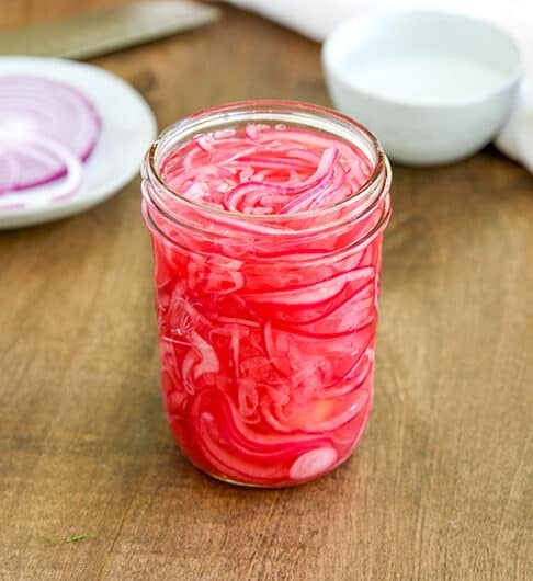 small jelly jar with thinly sliced red onions in it; back left is a small white plate with a slice of red onion; right hand side with white condiment bowl in a whole red onion on white cloth