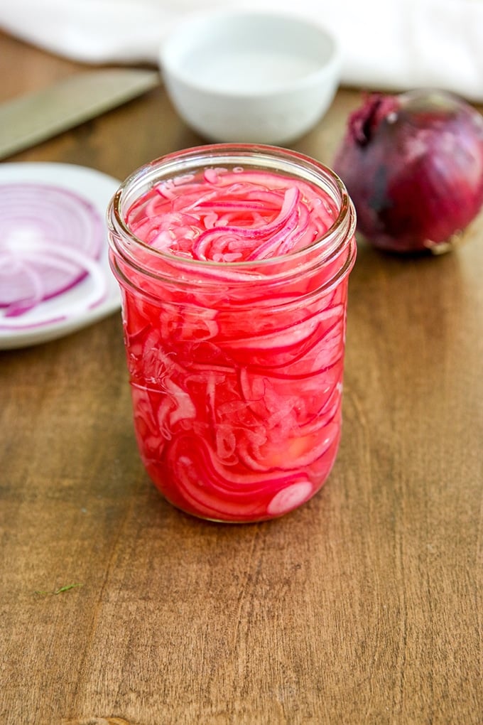 jar with pink thin slices of onion in a pickling liquid; red onion in background and small white plate with ring of red onion; white pinch bowl in background and white cloth