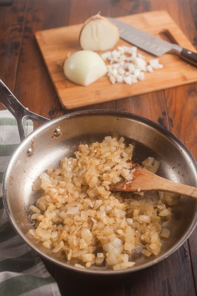stainless steel frying pan with sauteed chopped onions; wooden spatula in pan; white and green striped cloth in front left corner; cutting board in background with onion partially chopped and a knife