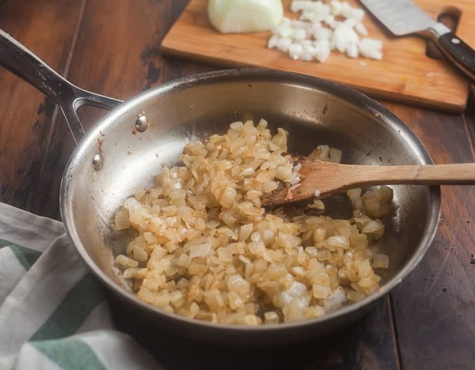 stainless steel frying pan with sauteed chopped onions; wooden spatula in pan; white and green striped cloth in front left corner; cutting board in background with onion partially chopped and a knife