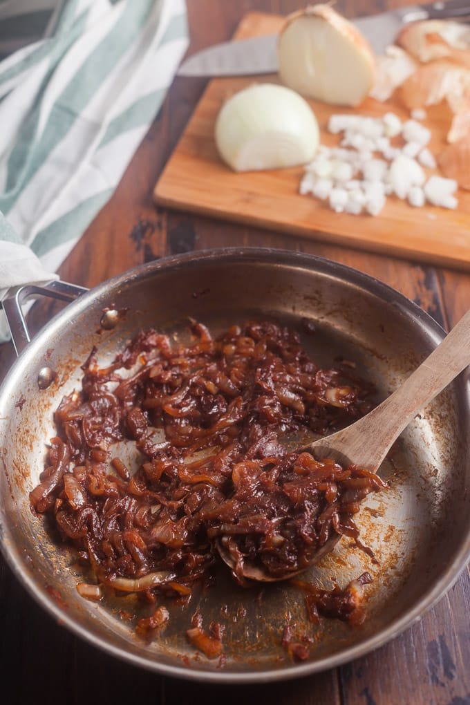 caramelized onions in a stainless steel frying pan with a wooden spoon in pan; white and green striped cloth in background; cuttiong board in background with partially chopped onion and knife