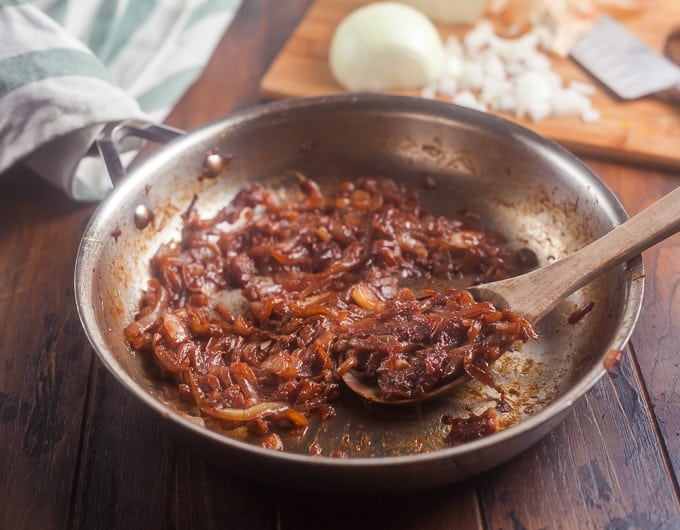 caramelized onions in a stainless steel frying pan with a wooden spoon in pan; white and green striped cloth in background; cuttiong board in background with partially chopped onion and knife
