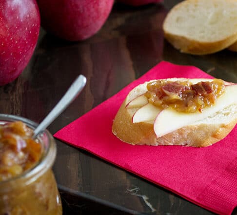 red paper napkin with baguette slice on top with 3 apple slices and a dollop of onion jam with bacon in it; 2 slices baguette in background, 2 red apples in back left corner and jar of onion jam in front corner with small spoon in it