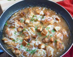 A large skillet with chicken and a brown sauce, topped with melted cheese, croutons, and parsley. There's a cutting board with a Vidalia onion and some parsley in the background.