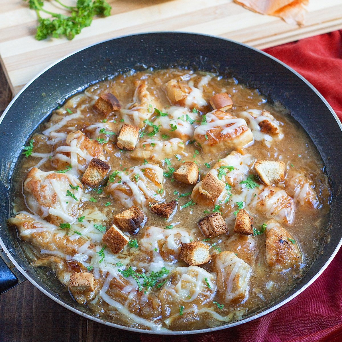 A large skillet with chicken and a brown sauce, topped with melted cheese, croutons, and parsley. There's a cutting board with a Vidalia onion and some parsley in the background.