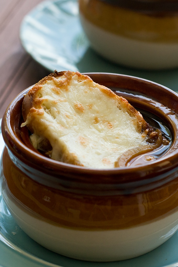 brown crock with french onion soup, toasted bread with melted cheese on top; white plate in background