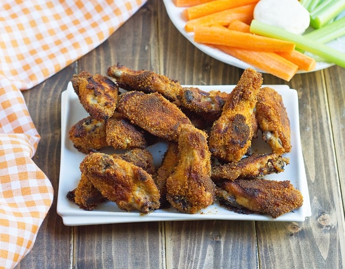 breaded cooked chicken wings on a white rectangle platter with an orange gingham cloth in the background and a plate with carrot and celery sticks