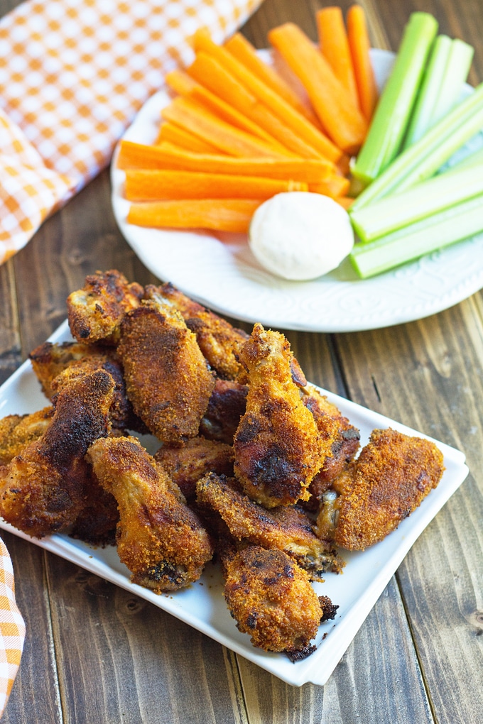 breaded cooked chicken wings on a white rectangle platter with an orange gingham cloth in the background and a plate with carrot and celery sticks
