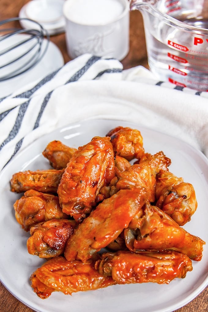buffalo chicken wings on a white plate; white cloth with blue stripes, pyrex measure with water in it, salt and whisk in background 