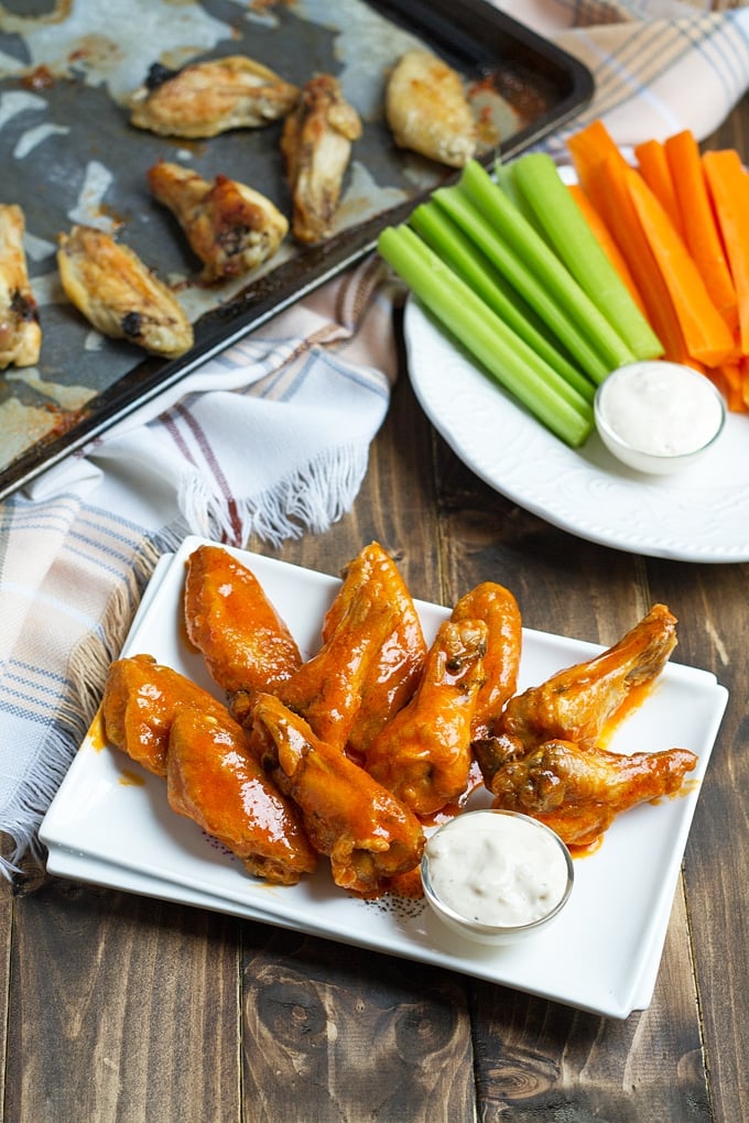 buffalo wings on rectangular white platter with blue cheese dressing in small bowl; white cloth with blue striping in back left; celery and carrots on plat in back right