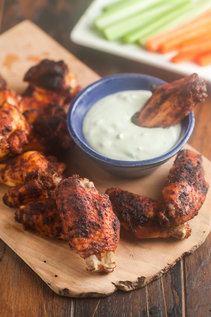 baked chicken wings with seasoning on the, around medium blue condiment bowl with blue cheese dressing in it and one wing in the dressing; all sitting on a brown cutting board