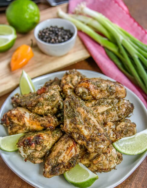 well seasoned chicken wings on light gray plate garnished with lime wedges; light pink cloth in background with bunch of green onions; cutting board with condiment bowl with seasonings, orange habanero pepper, and lime