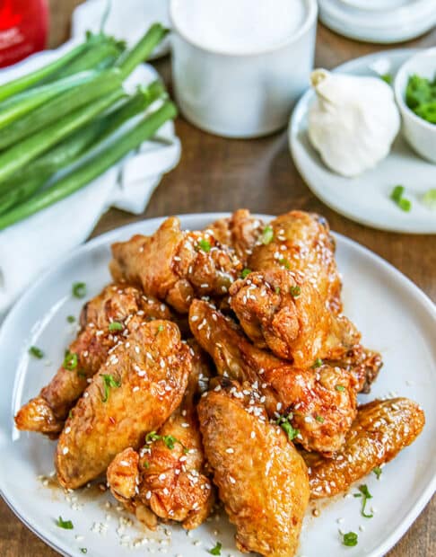 fried wings garnished with chive and sesame seeds on light gray plate; small condiment plate in background with garlic and chives on it; bunch of green onions in background